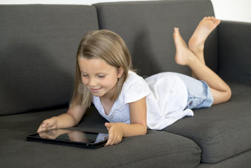 Les jeunes petites années douces et heureuses de la fille 6 ou 7 se trouvant sur le sofa à la maison de salon couchent à l'aide d photographie stock Les jeunes petites années douces et heureuses de la fille 6 ou 7 se trouvant sur le sofa à la maison de salon couchent à l'aide d photographie stock