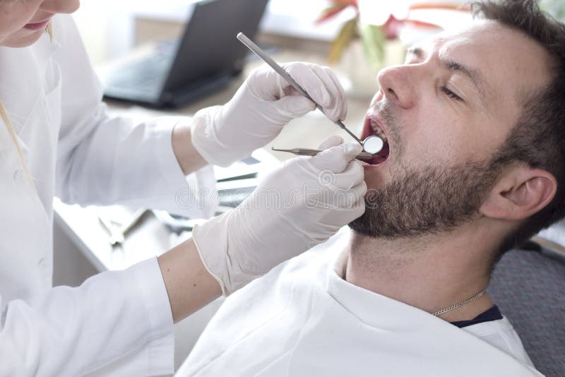 Les mains du dentiste féminin tiennent le miroir et l'excavatrice dentaires Un homme s'assied sur une chaise dentaire avec une bo