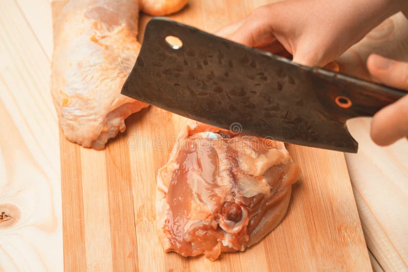 Female hands cut a chicken thigh at the junction of the bones with a chef`s cleaver. Female hands cut a chicken thigh at the junction of the bones with a chef`s cleaver.