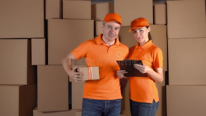 Les messagers dans l'uniforme orange se tenant contre le carton brun empile le backround Personnel de société de livraison image stock Les messagers dans l'uniforme orange se tenant contre le carton brun empile le backround Personnel de société de livraison image stock