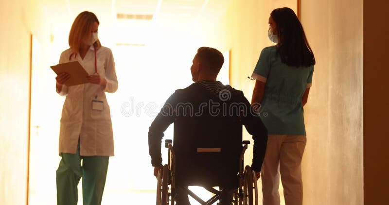 Les médecins accompagnent le patient en fauteuil roulant dans le couloir de l'hôpital banque de vidéos