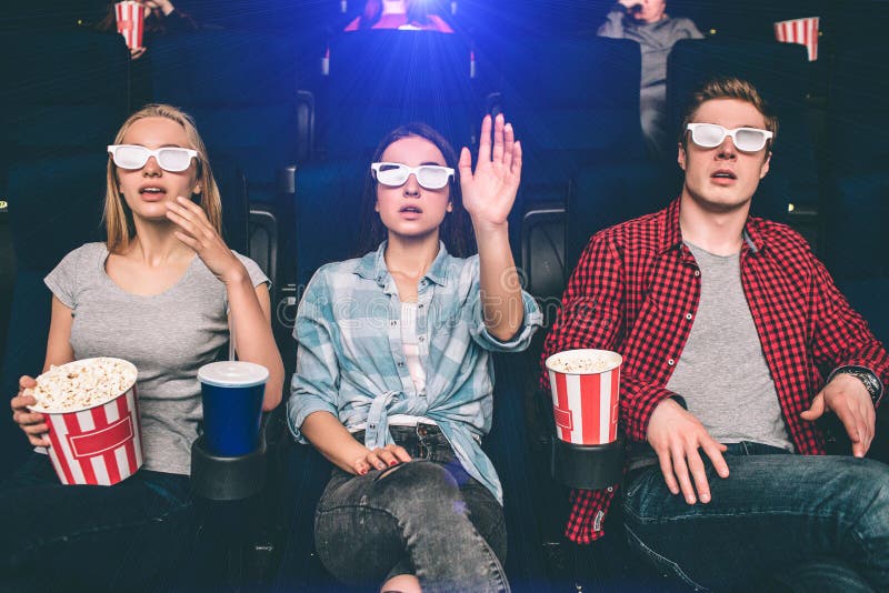Surprised and amazed people are sitting in chairs in cinema. They are watching movie and looking at the screen with glasses. Girl in the middle is showing her hand. Surprised and amazed people are sitting in chairs in cinema. They are watching movie and looking at the screen with glasses. Girl in the middle is showing her hand