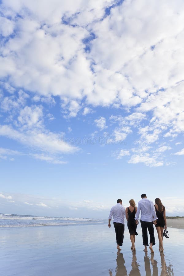 Four young people, two couples, holding hands, walking together on a deserted beach with a blue sky and clouds. Four young people, two couples, holding hands, walking together on a deserted beach with a blue sky and clouds.