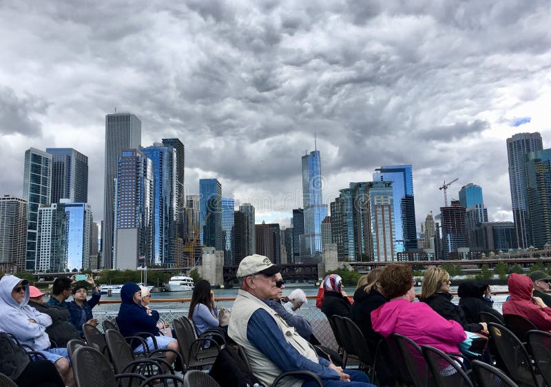 Storm clouds swirl above downtown Chicago, Illinois. Blue and gray buildings are lit up. Tourists in foreground ride an architecture tour boat. Storm clouds swirl above downtown Chicago, Illinois. Blue and gray buildings are lit up. Tourists in foreground ride an architecture tour boat.