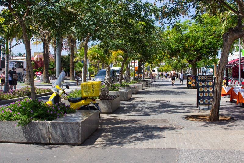 Tourists on a tree lined avenue with shops and cafes in Playa de Las Americas in the island of Teneriffe. The volcanic Teidi mountain range is in the background. Tourists on a tree lined avenue with shops and cafes in Playa de Las Americas in the island of Teneriffe. The volcanic Teidi mountain range is in the background