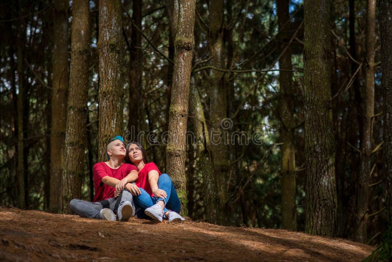 Lesbian Couple Sitting In The Woods Under A Tree With Sunlight On Them