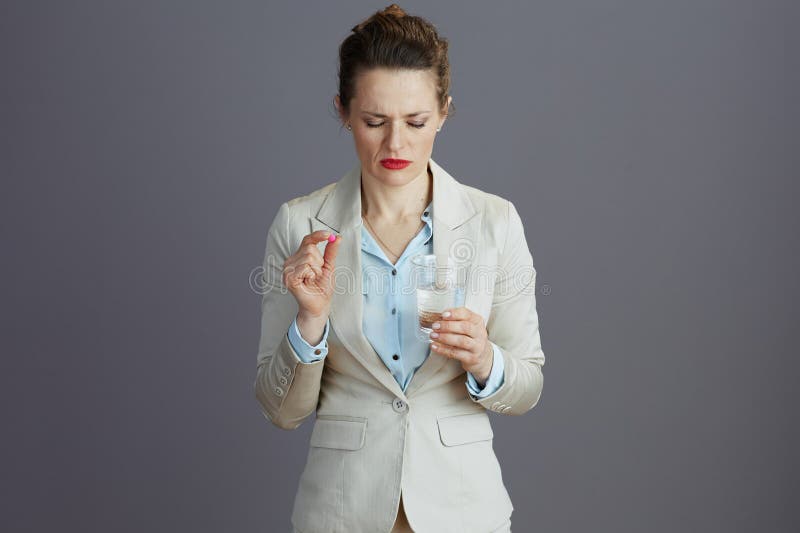 stressed 40 years old woman employee in a light business suit with glass of water and pill isolated on grey background. stressed 40 years old woman employee in a light business suit with glass of water and pill isolated on grey background