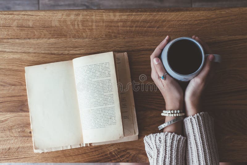 Person relaxing while reading a book and holding mug with hot morning coffee or tea on rustic wooden table in cafe. Warm sweater and boho jewelry on hands. Hygge weekend scene, top view. Person relaxing while reading a book and holding mug with hot morning coffee or tea on rustic wooden table in cafe. Warm sweater and boho jewelry on hands. Hygge weekend scene, top view.