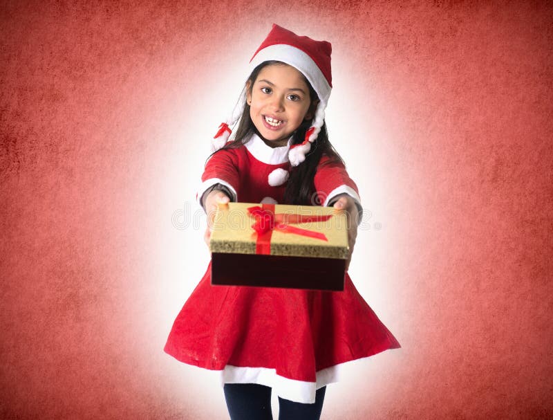 Cute Little Hispanic Girl in Santa Claus costume holding a Christmas Box Present as giving or receiving it on red Xmas studio background. Cute Little Hispanic Girl in Santa Claus costume holding a Christmas Box Present as giving or receiving it on red Xmas studio background