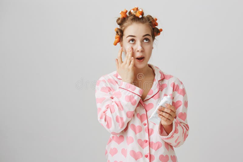 Cute feminine european girl wearing hair-curlers and pyjamas applying cream on clean skin, looking up and standing over gray background. Fashionable woman takes care of her appearance. Cute feminine european girl wearing hair-curlers and pyjamas applying cream on clean skin, looking up and standing over gray background. Fashionable woman takes care of her appearance.