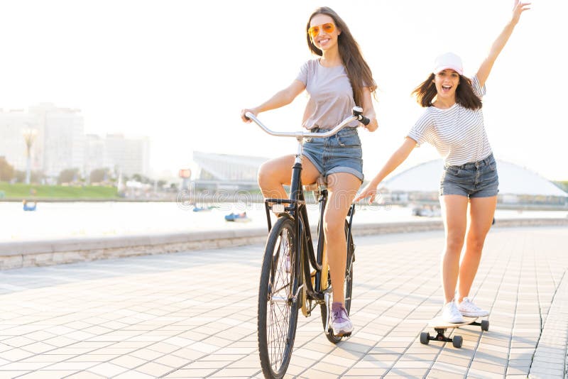 Leuke lachende jonge vrouw in jeansborrels op fiets die meisjesholding trekken terwijl het gebruiken van skateboard royalty-vrije stock foto's Leuke lachende jonge vrouw in jeansborrels op fiets die meisjesholding trekken terwijl het gebruiken van skateboard royalty-vrije stock foto's
