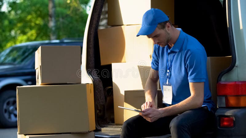 Leveringsarbeider die pakketbedrag in bestelwagen, inventarisatierapport controleren over tablet stock afbeelding Leveringsarbeider die pakketbedrag in bestelwagen, inventarisatierapport controleren over tablet stock afbeelding