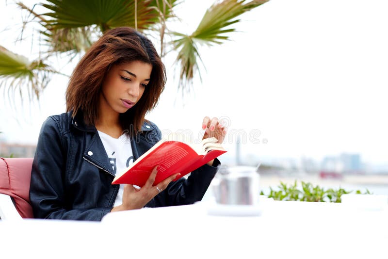 Attractive afro-american female reading fascinating book while sitting on the terrace of coffee shop, young brunette woman relaxing at weekend while read interesting book outdoors in cafe restaurant. Attractive afro-american female reading fascinating book while sitting on the terrace of coffee shop, young brunette woman relaxing at weekend while read interesting book outdoors in cafe restaurant