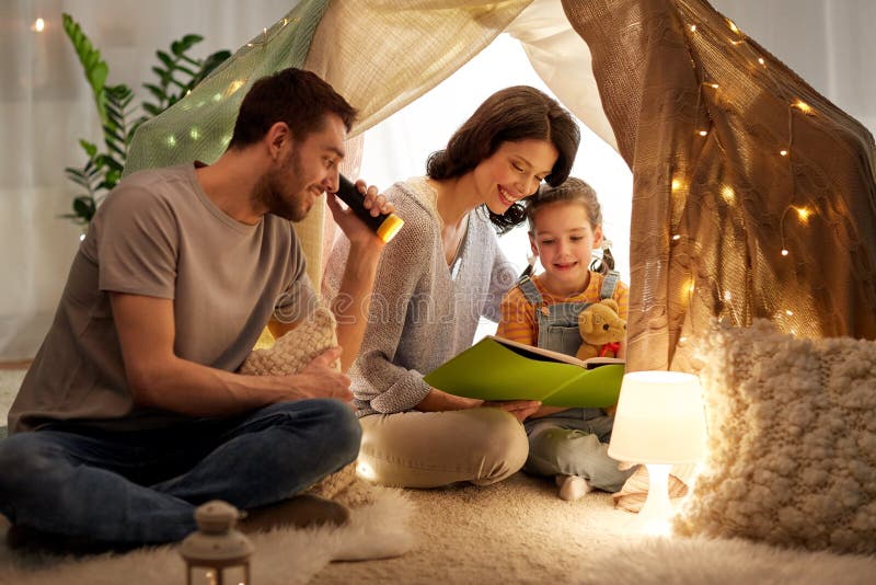 Family, hygge and people concept - happy mother, father and little daughter reading book with torch light in kids tent at night at home. Family, hygge and people concept - happy mother, father and little daughter reading book with torch light in kids tent at night at home