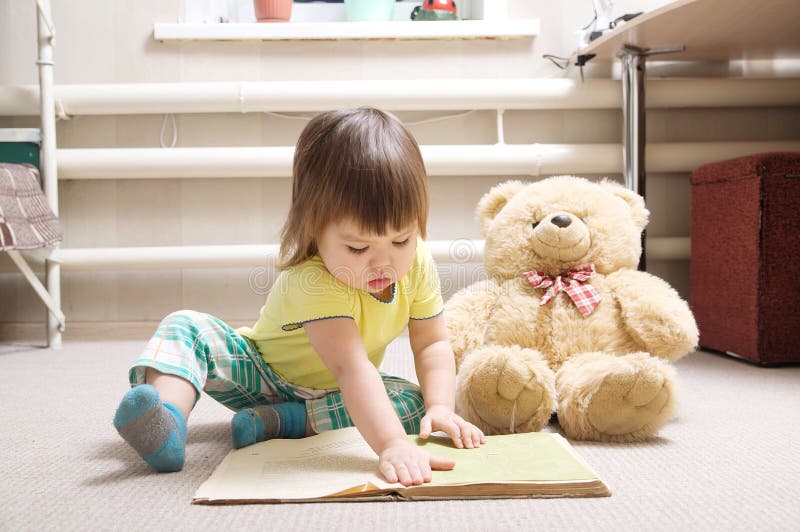 Libro di lettura della bambina dell'interno nella sua stanza su tappeto con l'orsacchiotto del giocattolo, bambino sveglio che gi immagini stock Libro di lettura della bambina dell'interno nella sua stanza su tappeto con l'orsacchiotto del giocattolo, bambino sveglio che gi immagini stock