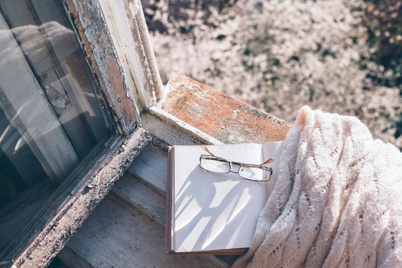 Opened book, cup of tea or coffee, glasses and knitted sweater on window sill over blooming tree view. Cozy spring weekend concept. The text on pages is not recognizable. Opened book, cup of tea or coffee, glasses and knitted sweater on window sill over blooming tree view. Cozy spring weekend concept. The text on pages is not recognizable