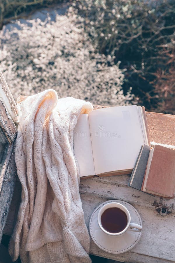 Opened book, cup of tea or coffee, glasses and knitted sweater on window sill over blooming tree view. Cozy spring weekend concept. The text on pages is not recognizable. Opened book, cup of tea or coffee, glasses and knitted sweater on window sill over blooming tree view. Cozy spring weekend concept. The text on pages is not recognizable