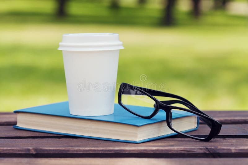 Libro, lentes y taza de papel con café en un banco en parque en un día soleado, leyendo en el verano, educación, libro de texto imágenes de archivo libres de regalías Libro, lentes y taza de papel con café en un banco en parque en un día soleado, leyendo en el verano, educación, libro de texto imágenes de archivo libres de regalías