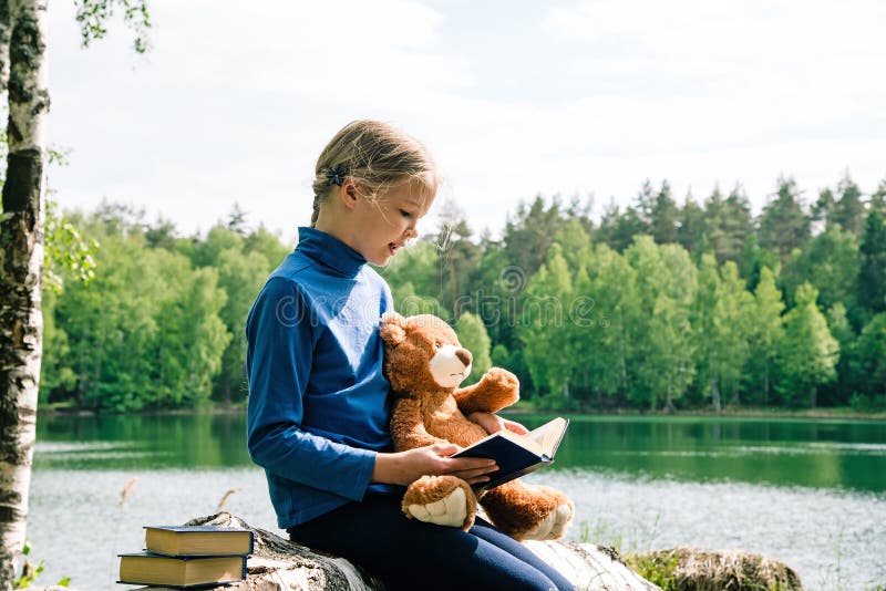 Libro sveglio dell'orsacchiotto della lettura della ragazza al picnic Migliori amici che si siedono sull'albero caduto del lago d immagine stock libera da diritti Libro sveglio dell'orsacchiotto della lettura della ragazza al picnic Migliori amici che si siedono sull'albero caduto del lago d immagine stock libera da diritti