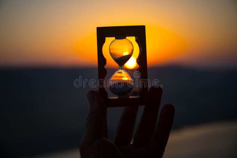 Life time passing concept. Hand holding hourglass with sunset sky background.