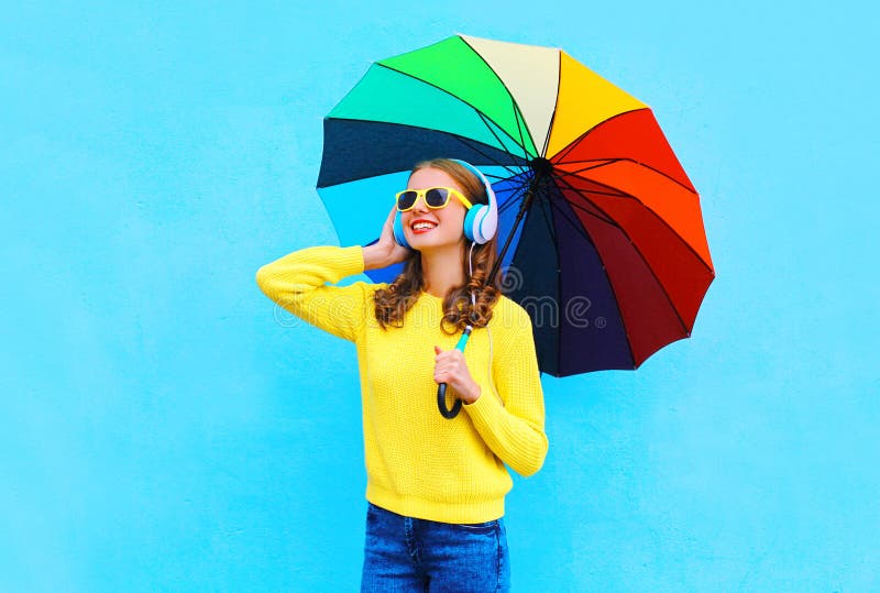 Lifestyle portrait smiling young woman listens to music in headphones with colorful umbrella in autumn day over colorful blue