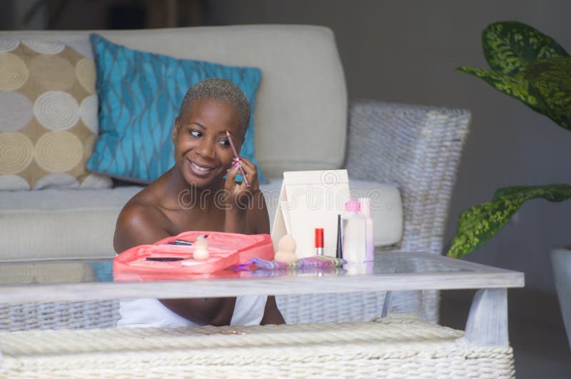 Lifestyle portrait of young happy and beautiful black afro American woman wrapped in towel applying face makeup eyebrow profiling pencil looking herself in the mirror at home living room