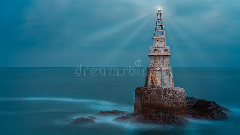 Old white lighthouse tower isolated in calm blue sea water after sunset. Landscape of lighthouse emitting light and rays
