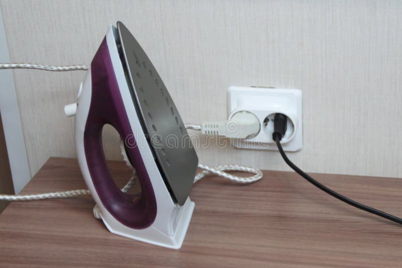 A Lilac Electric Iron Stands on a Table Plugged into a Wall Outlet