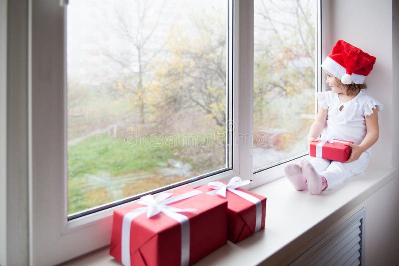 Lilla flickan i jultomtenhatt sitter på fönsterfönsterbräda och den hållande gåvan royaltyfria foton Lilla flickan i jultomtenhatt sitter på fönsterfönsterbräda och den hållande gåvan royaltyfria foton