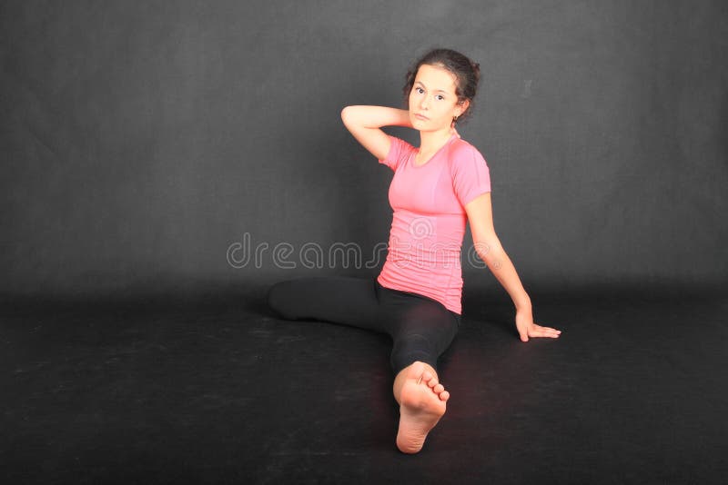 Pretty girl - barefoot brunette schoolkid dressed in pink t-shirt and black leggings exercising stretching while sitting on black background. Active childhood concept. Pretty girl - barefoot brunette schoolkid dressed in pink t-shirt and black leggings exercising stretching while sitting on black background. Active childhood concept.