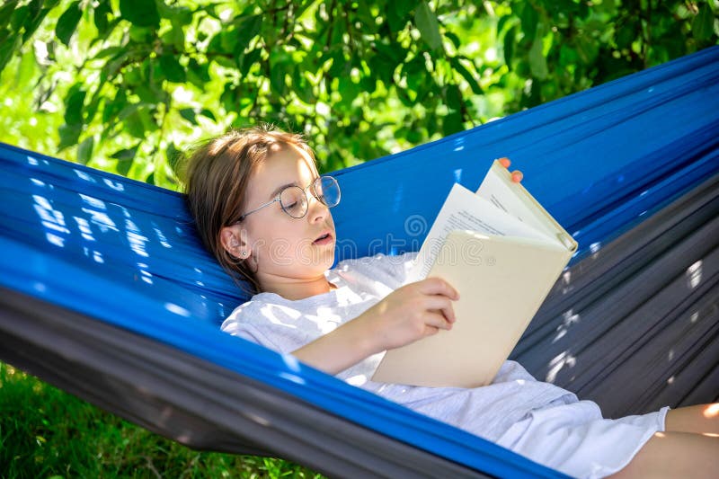 Cute teen girl in glasses reads a book while lying in a hammock in the garden. Cute teen girl in glasses reads a book while lying in a hammock in the garden.