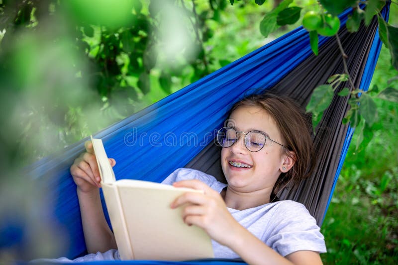 Cute teen girl in glasses reads a book while lying in a hammock in the garden. Cute teen girl in glasses reads a book while lying in a hammock in the garden.