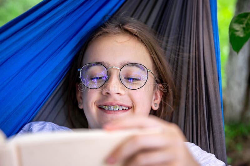 Cute teen girl in glasses reads a book while lying in a hammock in the garden. Cute teen girl in glasses reads a book while lying in a hammock in the garden.
