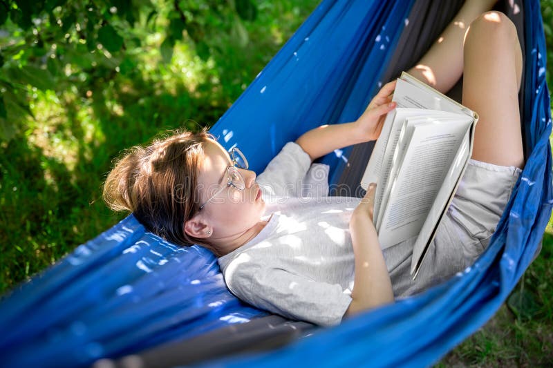 Cute teen girl in glasses reads a book while lying in a hammock in the garden. Cute teen girl in glasses reads a book while lying in a hammock in the garden.