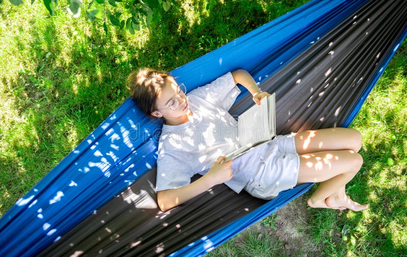 Cute teen girl in glasses reads a book while lying in a hammock in the garden, top view. Cute teen girl in glasses reads a book while lying in a hammock in the garden, top view.