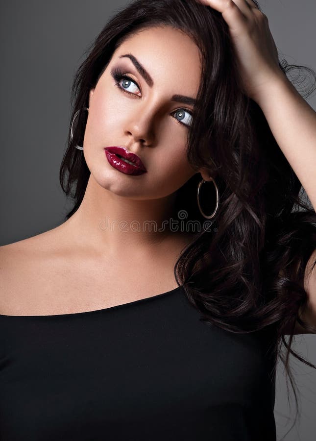 Beautiful bright makeup blue eyes woman with long black curly volume black hair style, burgundy lipstick with vamp looking up on grey background. Closeup. Beautiful bright makeup blue eyes woman with long black curly volume black hair style, burgundy lipstick with vamp looking up on grey background. Closeup