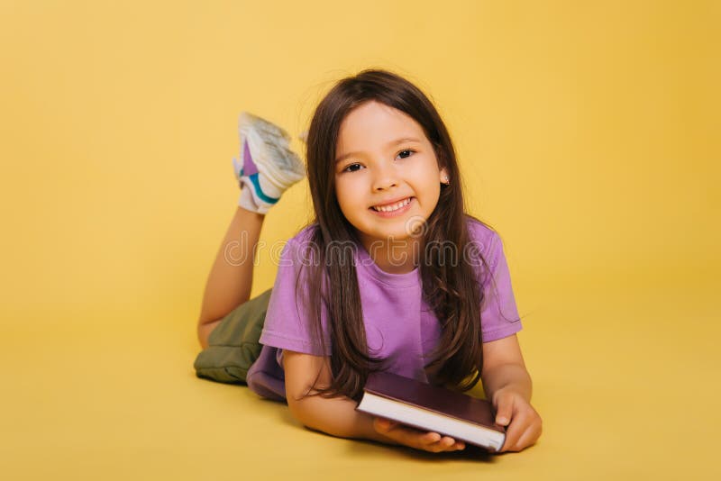 beautiful little girl reads a book while lying. Cute baby teaches lessons on a yellow background. Photo in studio. beautiful little girl reads a book while lying. Cute baby teaches lessons on a yellow background. Photo in studio