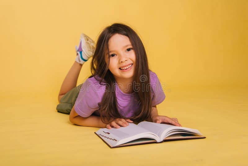 beautiful little girl reads a book while lying. Cute baby teaches lessons on a yellow background. Photo in studio. beautiful little girl reads a book while lying. Cute baby teaches lessons on a yellow background. Photo in studio