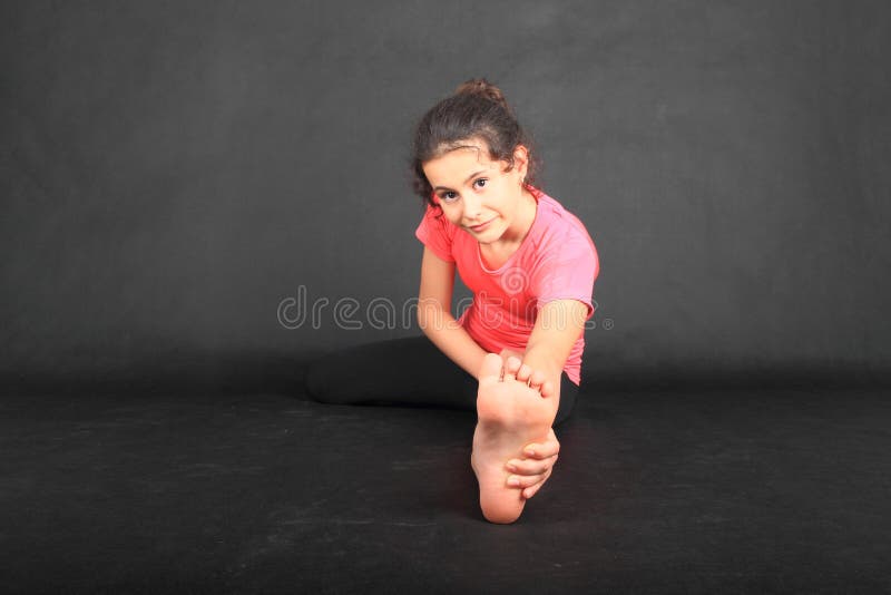 Pretty girl - barefoot brunette schoolkid dressed in pink t-shirt and black leggings exercising stretching - holding sole of her bare foot while sitting on black background. Active childhood concept. Pretty girl - barefoot brunette schoolkid dressed in pink t-shirt and black leggings exercising stretching - holding sole of her bare foot while sitting on black background. Active childhood concept.