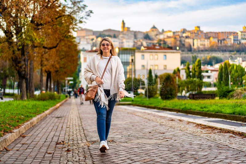 Linda mulher de cabelos longos de meia-idade usando jeans suéter bege óculos de sol e bolsa de couro andando fotografia de stock
