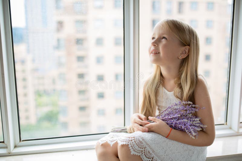 Cute little caucasian girl with blonde hair holding bouquet of purple flowers at home. Kid standing near window during coronavirus covid-19 pandemic quarantine. High quality photo. Cute little caucasian girl with blonde hair holding bouquet of purple flowers at home. Kid standing near window during coronavirus covid-19 pandemic quarantine. High quality photo