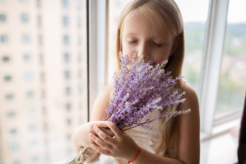 Cute little caucasian girl with blonde hair holding bouquet of purple flowers at home. Kid standing near window during coronavirus covid-19 pandemic quarantine. High quality photo. Cute little caucasian girl with blonde hair holding bouquet of purple flowers at home. Kid standing near window during coronavirus covid-19 pandemic quarantine. High quality photo