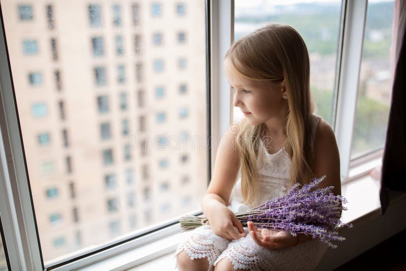 Cute little caucasian girl with blonde hair holding bouquet of purple flowers at home. Kid standing near window during coronavirus covid-19 pandemic quarantine. High quality photo. Cute little caucasian girl with blonde hair holding bouquet of purple flowers at home. Kid standing near window during coronavirus covid-19 pandemic quarantine. High quality photo