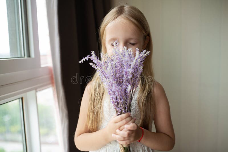 Cute little caucasian girl with blonde hair holding bouquet of purple flowers at home. Kid standing near window during coronavirus covid-19 pandemic quarantine. High quality photo. Cute little caucasian girl with blonde hair holding bouquet of purple flowers at home. Kid standing near window during coronavirus covid-19 pandemic quarantine. High quality photo