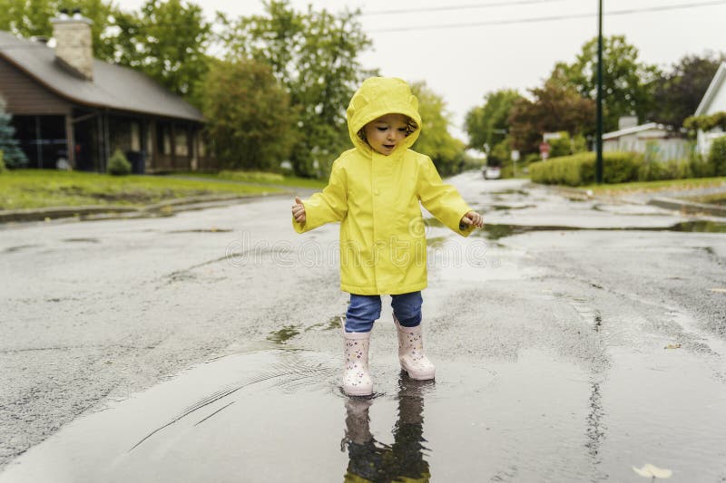 Una linda niña graciosa con un abrigo impermeable amarillo y botas jugando en la lluvia