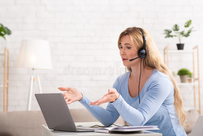 Online business meeting. Adult woman with headset communicates with client on laptop at workplace at home. Online business meeting. Adult woman with headset communicates with client on laptop at workplace at home