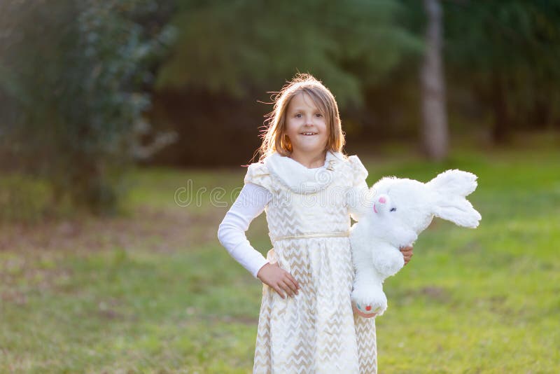 Liten Caucasian flicka i festlig klänning med den flotta vita leksakkaninen och att se till kameran som ler royaltyfri foto Liten Caucasian flicka i festlig klänning med den flotta vita leksakkaninen och att se till kameran som ler royaltyfri foto