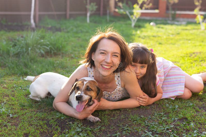 Liten flicka i rosa klänning med mamma och Jack Russell skräckhund som lägger på gräs. Titta på kameran och le royaltyfri foto