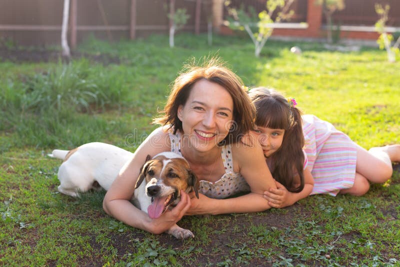 Liten flicka i rosa klänning med mamma och Jack Russell skräckhund som lägger på gräs. Titta på kameran och le arkivfoton