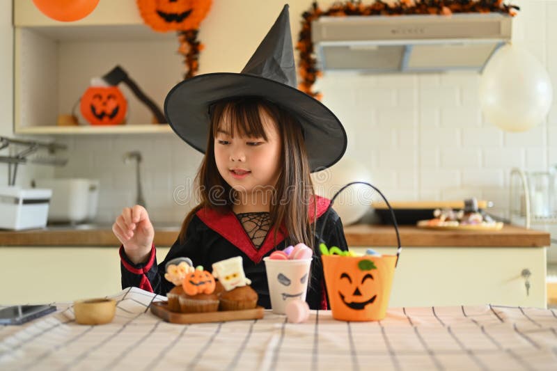 Liten flicka utklädd som en häxa som dekorerar halloween cupcakes med olika monster på köksbord royaltyfri bild